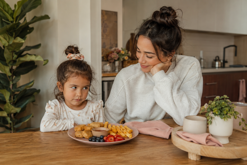 A child cautiously trying kid-friendly plant-based food swaps with meatless nuggets, fries, fruit, and dipping sauce at the kitchen table.