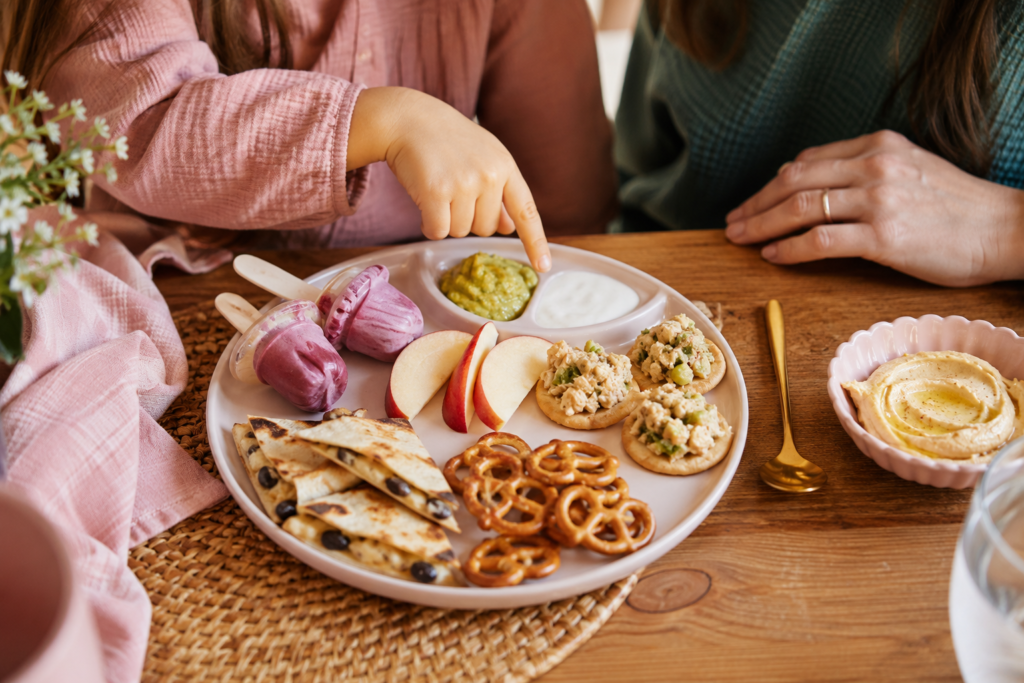  Kid-friendly plant-based tasting plate with mini bean quesadillas, chickpea salad crackers, smoothie popsicles, fruit, pretzels, and hummus.