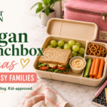 Vegan lunchbox with a sandwich, grapes, carrots, cucumber, hummus, and pretzels beside a pink water bottle and lunch bag.